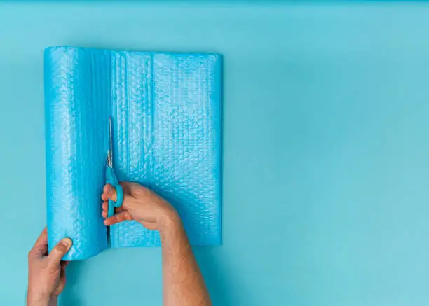 Overhead view of caucasian adult male hands packing with blue bubblewrap and cutting with scissors. Blue background with room for copy space.