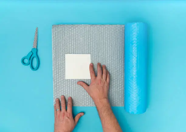 Overhead view of caucasian adult male hands packaging a blank white square paper with bubble wrap and scissors nearby. Blue background with room for copy space.