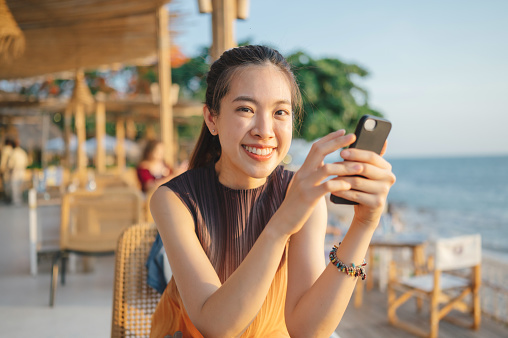 Solo Cheerful Asian women enjoy sunset scenics using smart phone enjoy social media at tropical beach sea sit on beach chair
