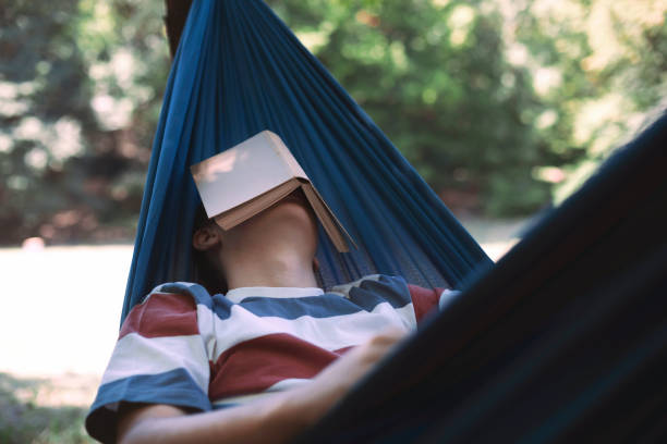 Mid adult woman laying relaxed on hammock in a public park at sunset. She is browsing the web or taking photos with a digital tablet.