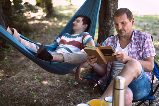 Young male teenager sleeping on hammock while his father reading a book. Camping. Reading a book. Resting. Relaxation.