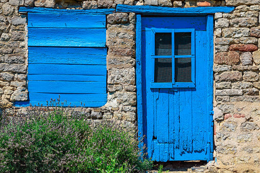 A very blue and very old door and window are glowing in the very old stone wall of a barn.
