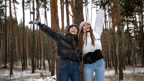 Two laughing teenage girls cheering and having fun because of snow in forest. People playing outdoors, winter holidays and vacation, active leisure