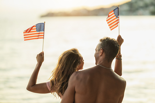 Rear view of a loving couple with American national flag enjoying a relaxing day on the beach.