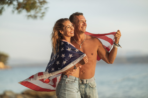 A smiling couple with American national flag enjoying a relaxing day on the beach.