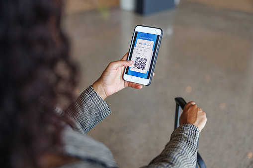 Close-up shot of a Latin-American business woman looking attentive at her digital boarding pass on her phone to check her departure date and wait for her upcoming flight, surrounded with other passengers.