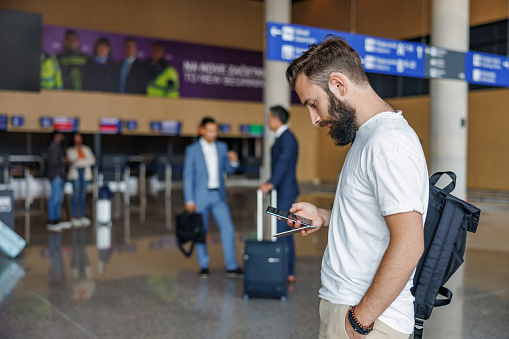 Three-quarter side shot with blurred background of a mid-adult Caucasian passenger looking attentive to his cellphone and the screen of the departure gates of the airport to make sure he goes to the right direction for his upcoming trip.