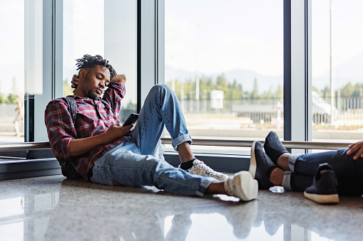 Full length side shot with blurred background of a mid-adult Latin-American man comfy lying on the airport floor next to a big window while waiting to be called for the flight.