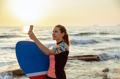 A young Caucasian woman is standing on the beach and taking a picture of the horizon using her phone. She's holding a skimboard.