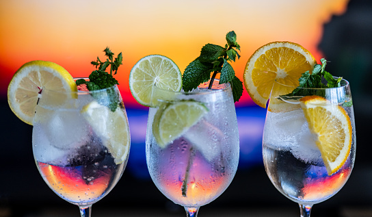 Three Colorful Gin Tonic Cocktails In Glasses At Bar Counter In Pub Or Restaurant With Out Of Focus Background Of A Sunset On A Beach