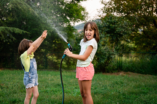 Girls splashing water from a hose in the backyard, in the garden, summer fun on a hot day and a happy childhood