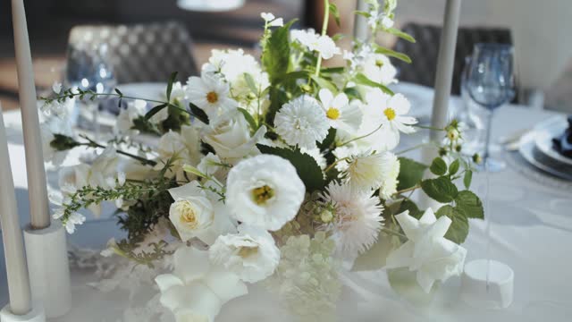 Dolly shot of wedding table decoration with bouquets of fresh white tender flowers with greenery, wine glasses on the background camera moving slow motion.