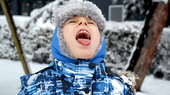 Funny little boy in winter hat cathcing snowflakes with tongue in park during snow storm.