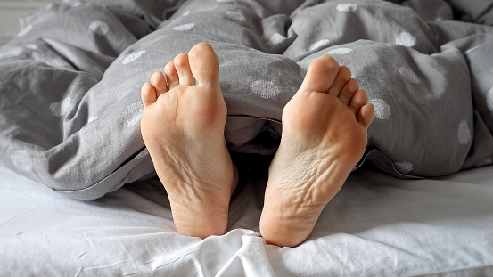 Closeup of a pair of feet belonging to a woman, showing from under the blankets on a bed. A beautiful depiction of the joys of unwinding at home
