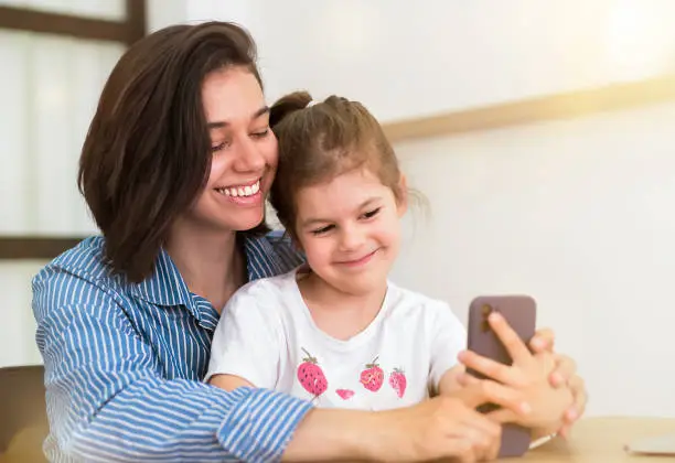 Photo of Happy family, cute little kid, daughter with mom, laughing, using smart phone, watching funny video on social media app, taking selfie, looking at mobile phone screen at home