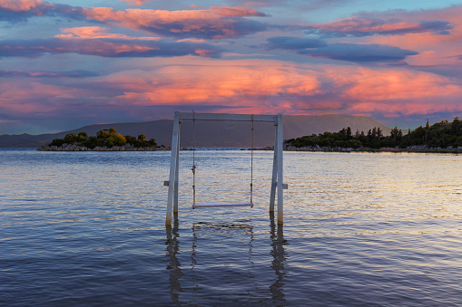 A white wooden swing standing on the edge of a shallow sea, a green island in the background and a dramatic sky with clouds