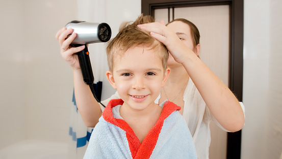 Portrait of mother and little boy drying hair with hairdryer after having bath. Concept of child hygiene and health care at home. Caring parents and kids at home.