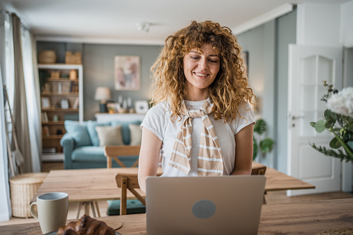 A young woman full of enthusiasm and happiness is working on her laptop in the comfort of her new home. Her smiling and cheerful expression clearly speaks of her satisfaction and excitement as she devotes herself to her work.