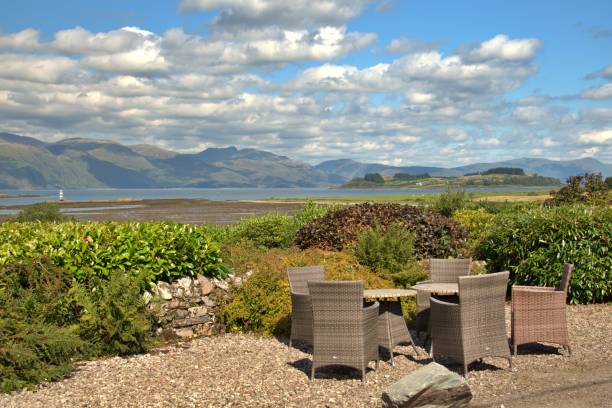 Panoramic view of Scottish highlands from village Appin on the west coast of Scotland,  table with some chairs on the side of the road,  white lighthouse visible in a distance