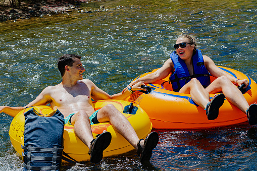 Golden, Colorado, USA, August 12, 2023, Two river tubers in orange and yellow river tubes, male and female , floating in the calm waters  of Clear Creek holding hands