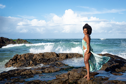 Beautiful smiling woman with black power hair wearing light green outfit with white bikini standing on the rocks of a beach. Blue sky and clouds in the background. Rio Vermelho Beach, Salvador, Brazil.