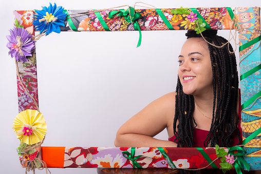 Photo of a beautiful woman with braids in her hair behind a frame decorated with flowers and colorful cloths in a horizontal position. White color background.