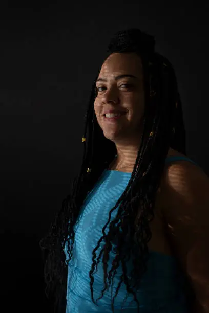 Half-closed portrait of a pretty woman in profile wearing blue outfit with braids in her hair and geometric shadows on her face. Studio portrait with black background.