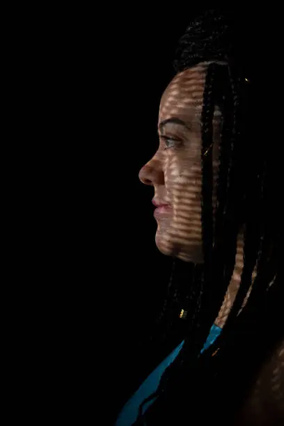 Half-closed portrait of a pretty woman in profile wearing blue outfit with braids in her hair and geometric shadows on her face. Studio portrait with black background.