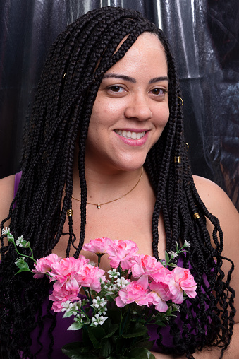 Portrait of a beautiful woman with braids in her hair wearing a lilac outfit holding a bouquet of flowers looking at the camera. Isolated on silver color background. Positive and happy person.