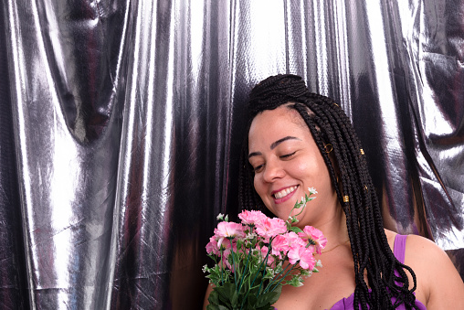 Portrait of a beautiful woman with braids in her hair wearing a lilac-colored outfit holding a bouquet of flowers. Isolated on silver color background. Positive and happy person.