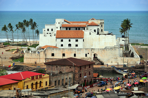 Elmina, Ghana: Benya Street and São Jorge da Mina Castle, also known simply as Mina or 'Feitoria da Mina', built by Portugal in 1482, the first trading post built on the Gulf of Guinea, and the oldest European building in existence south of the Sahara. Later also used by the Dutch to buy slaves from their African partners for their clients in the Americas - colonial architecture - Atlantic Ocean in the background - UNESCO World Heritage Site.