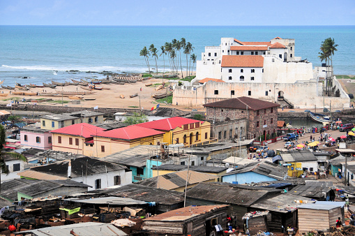 Elmina, Ghana: view from Saint Jago hill towards the town and São Jorge da Mina Castle, also known simply as Mina or 'Feitoria da Mina', built by Portugal in 1482. First established as a trade settlement, the castle later became one of the most important stops on the route of the Atlantic slave trade. The Dutch seized the fort from the Portuguese in 1637. The slave trade operated under the Dutch until 1814. In 1872, the Dutch Gold Coast, including the fort, became a British possession - colonial architecture - Gulf of Guinea / Atlantic Ocean in the background - UNESCO World Heritage Site.