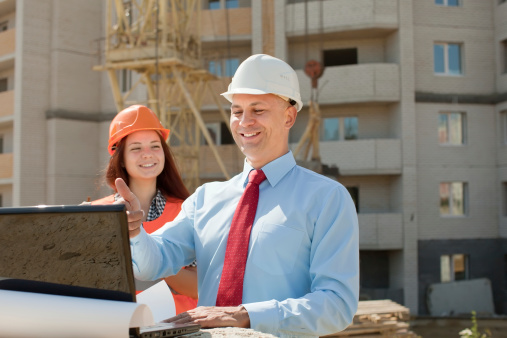 Young asian male engineers, wearing gray suits, yellow helmets and drawings in their hands, work on the construction site.