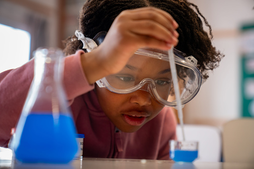 Black 8 year old girl in science class looking very focused while using a dripper with a blue liquid wearing protective goggles - School life concepts