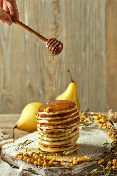 Classic American pancakes stacked on a pale wooden platter with sprigs of sea buckthorn and a pair of yellow pears. Honey flows from a wooden spoon. Recipe Blog Post. Serving a dish in a restaurant. panqueques americanos clásicos apilados en un plato de madera pálida con ramitas de espino cerval de mar y un par de peras amarillas. la miel fluye de una cuchara de madera. publicación del blog de recetas. servir un plato en un restaurante.