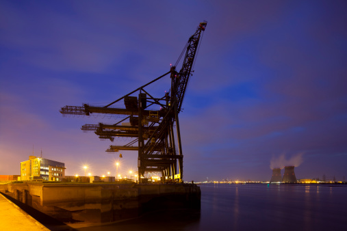 A container terminal with tall cranes with a nuclear power station in the background. Night shot taken in Antwerp, Belgium.