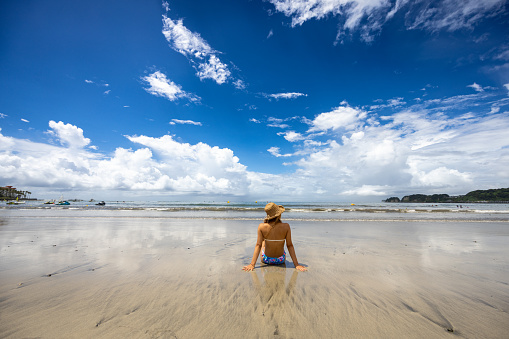 Rear view of tanned woman in bikini sitting in summer beach - wide shot