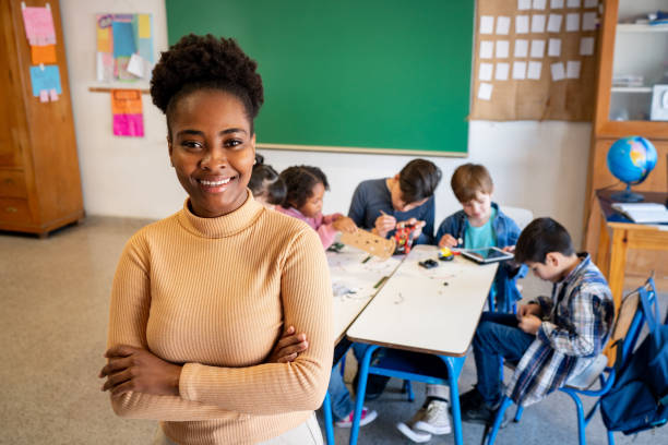 istock Black female teacher with arms crossed and smiling at the camera while her students work focused on their project during robotics class 1628348763