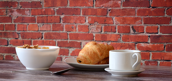A plate of oatmeal with nuts, two fresh croissants and a cup of black coffee on a table against a brick wall. Close-up.