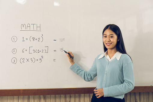 Asian female teacher is teaching students at the classroom while pointing at numbers on the white board.