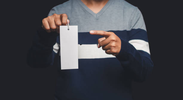 istock Hand holding and pointing at a blank white label or price tag while standing on a black background 1628579451