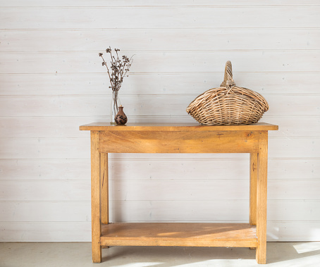 Oak side table with accessories including seedpod, dried twigs and wicker basket against white painted timber wall (selective focus)