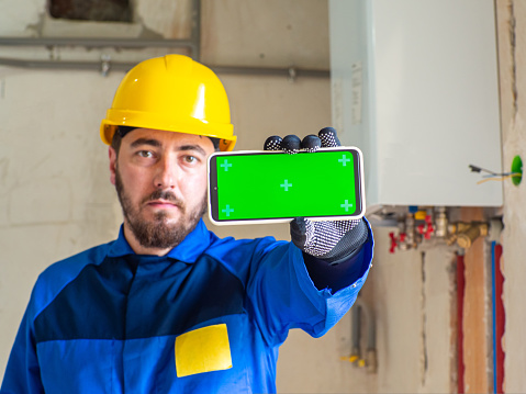 Portrait of worker or engineer in blue work clothes and yellow safety helmet with space for text. A man at a construction site holding a phone with chroma key screen in his hand and showing it.