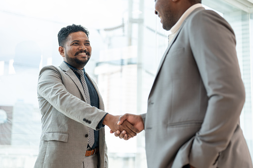 A professional and smiling Indian-Asian businessman shakes hands with an African American business partner after a meeting. Greeting, welcome, congratulations, successful business dealing, partnership