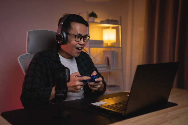 istock Asian influencer man wearing glasses smile youtuber using joystick playing game live streaming greeting video conferrence with laptop and wearing headphone. Asian man teaching online concept. 1628721571