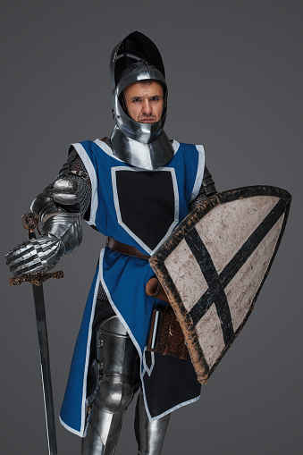Valiant medieval warrior wearing armor and blue surcoat with raised visor, gripping his sword, bravely staring into the camera, set against a gray backdrop