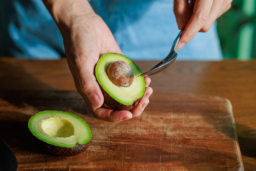 A close-up photograph capturing a woman skillfully peeling an avocado with a spoon on a cutting board, with the beautiful morning sun shining on the avocado.