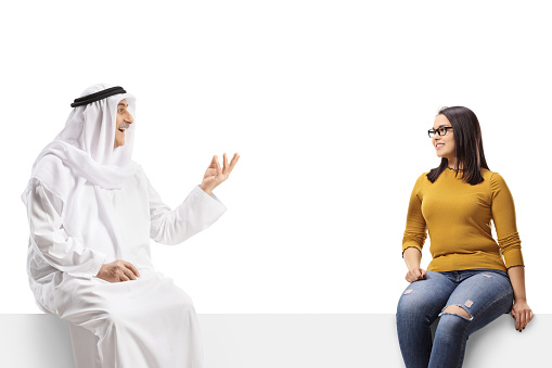 Arab man in a robe sitting on a blank panel and talking to a young woman isolated on white background
