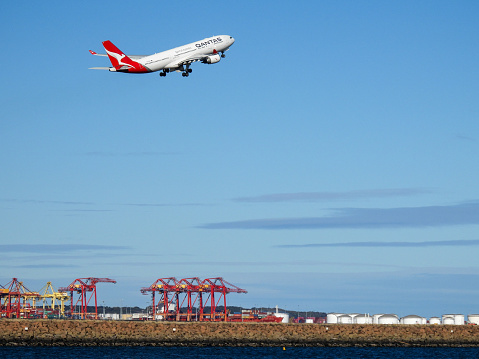 A Qantas Airbus A330-202 plane, registration VH-EBM, has taken off to the south of Sydney Kingsford-Smith Airport and heading to Manila as flight QF19.  In the distance are cranes of the Port Botany Container Terminal and fuel storage tanks at Kurnell. This image was taken from Botany Bay, Kyeemagh on a windy, cold and sunny afternoon on 19 August 2023.