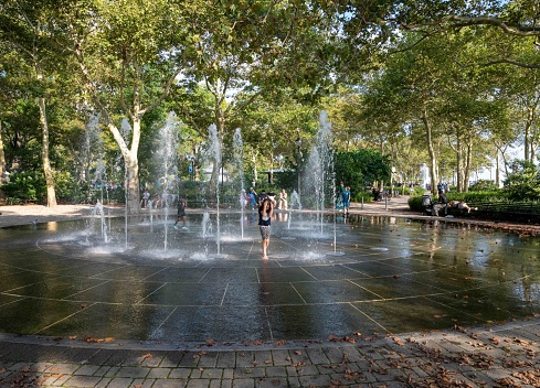New York, NY, USA 08-14-2023 Childeren splash and play in a public New York City fountain at Battery Park in Manhattan. People sitting on park benches under shade trees background. Editorial use only